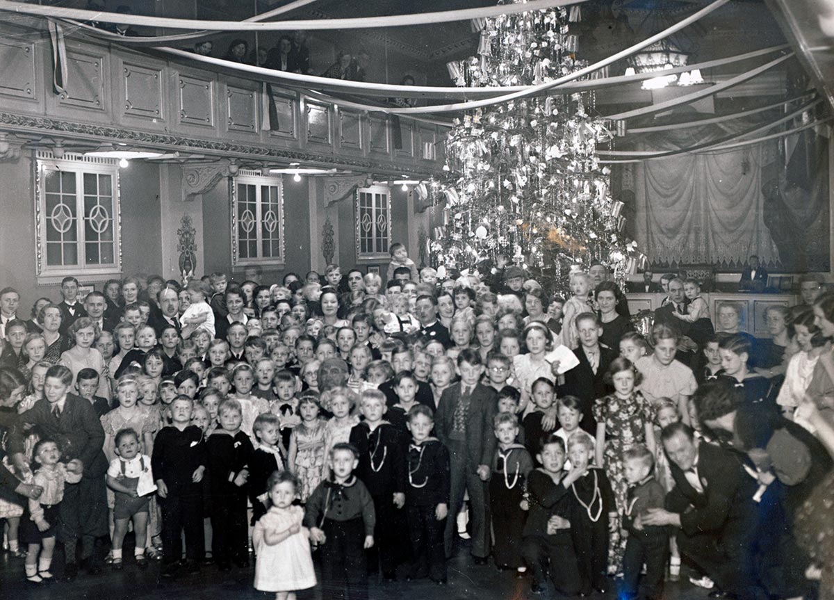 Festklædte medlemmer af Familieklubben har her taget opstilling foran juletræet i festsalen i Gimle. Foto: Ukendt fotograf, Københavns Stadsarkiv