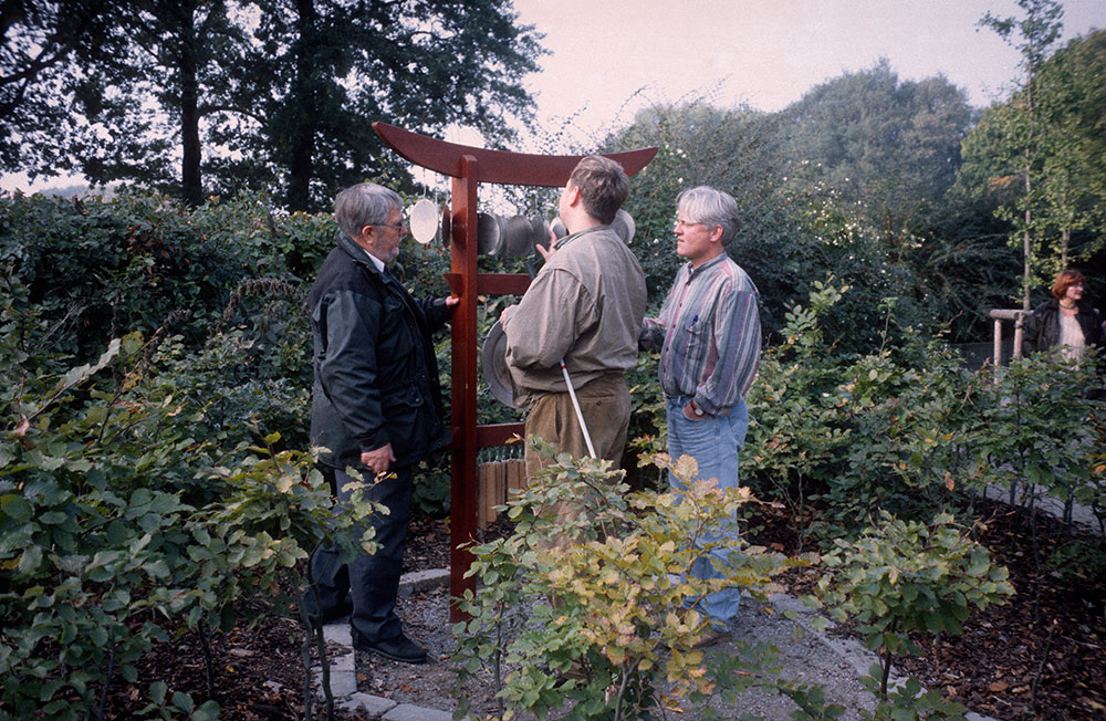 Tre persone står i have foran et vindspil med forskellige plader af træ og metal.