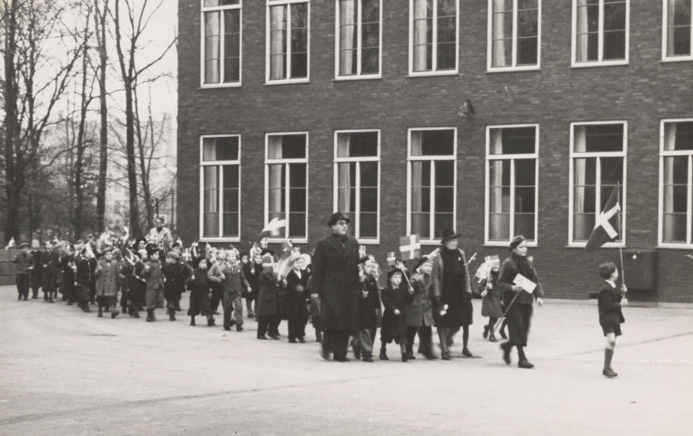 Børn med flag går på række ind i skolegård.