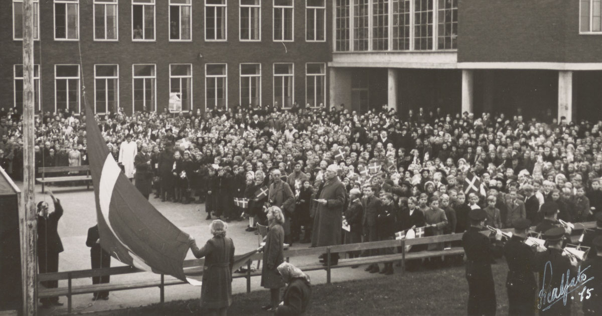 Forsamling af børn og voksne i skolegård med flag. Forsamlingen vender mod flagstang, hvor Dannebrog bliver hejst.