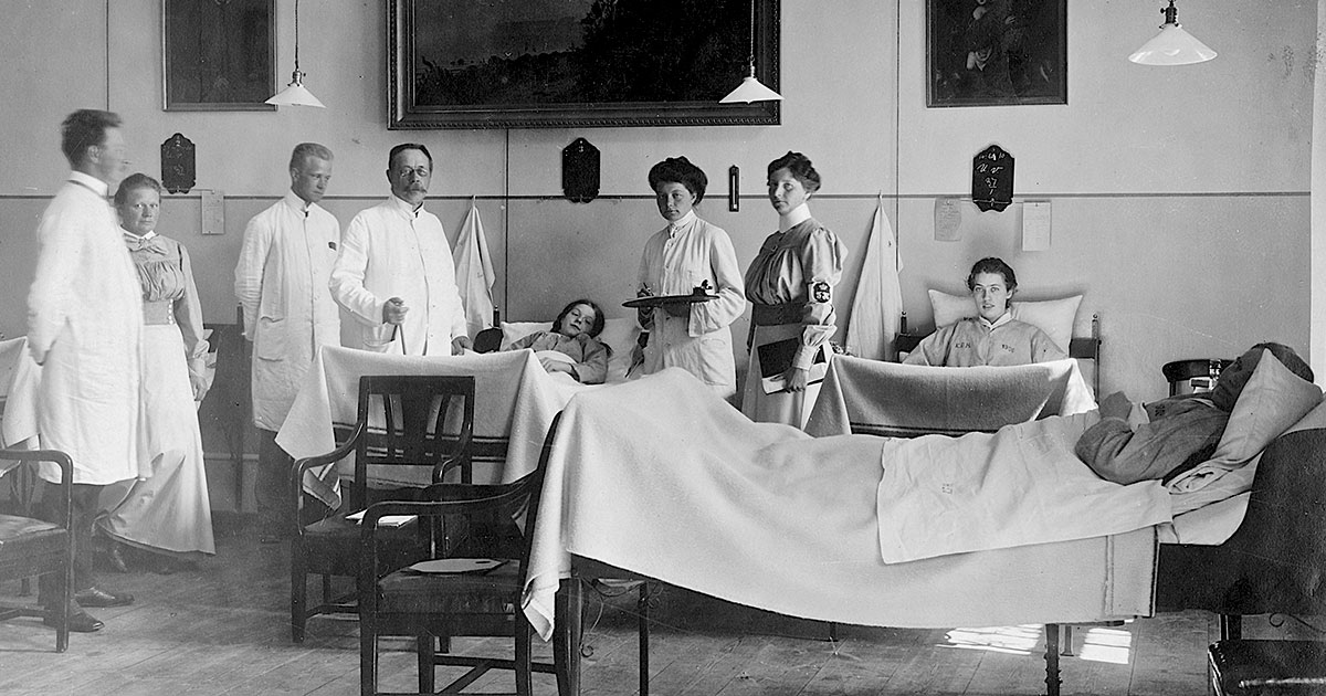 Hospitalsstue med tre liggende patienter i senge. På stuen står tre mænd og en kvinde i hvide kitler samt to kvinder med hvide forklæder.