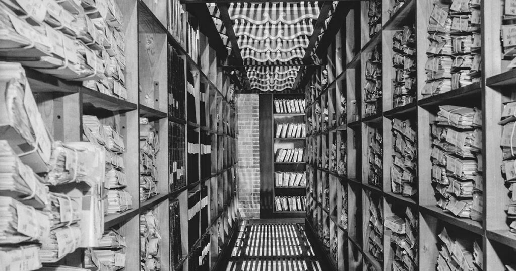 Arkivkælder med ristegulv, murstensendevæg og reoler fra gulv til loft. På reolerne ligger bundter af dokumenter og protokoller.