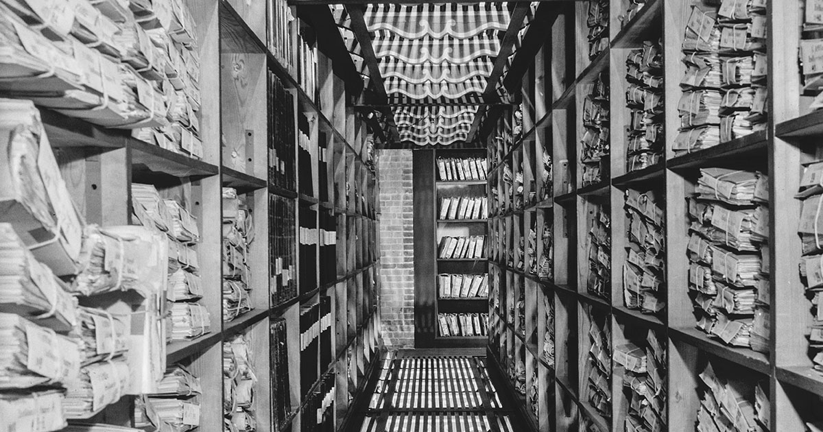 Arkivkælder med ristegulv, murstensendevæg og reoler fra gulv til loft. På reolerne ligger bundter af dokumenter og protokoller.