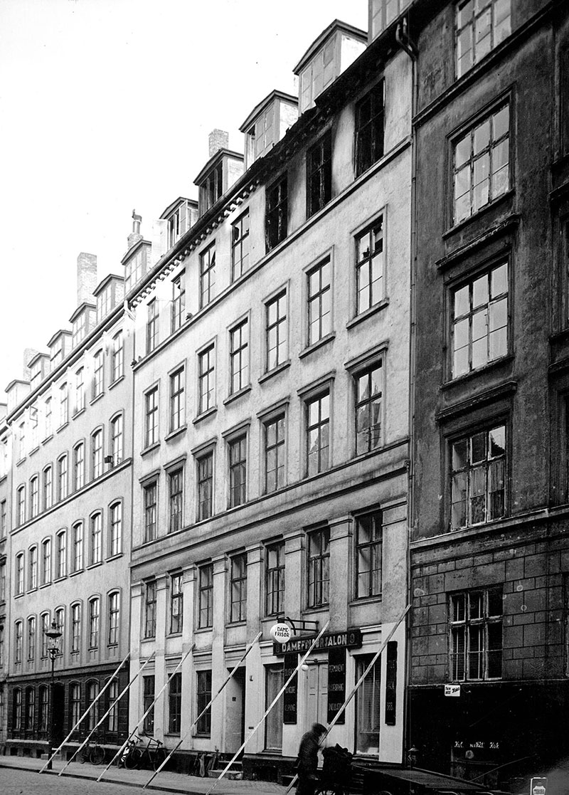 Gadeforløb med lejlighedsfacader. Øverst ses tre tilsodede vinduer. Nederst ses facaden på en damefrisørsalon og en kulforretning. I forgrunden ses en mand ved et lad.