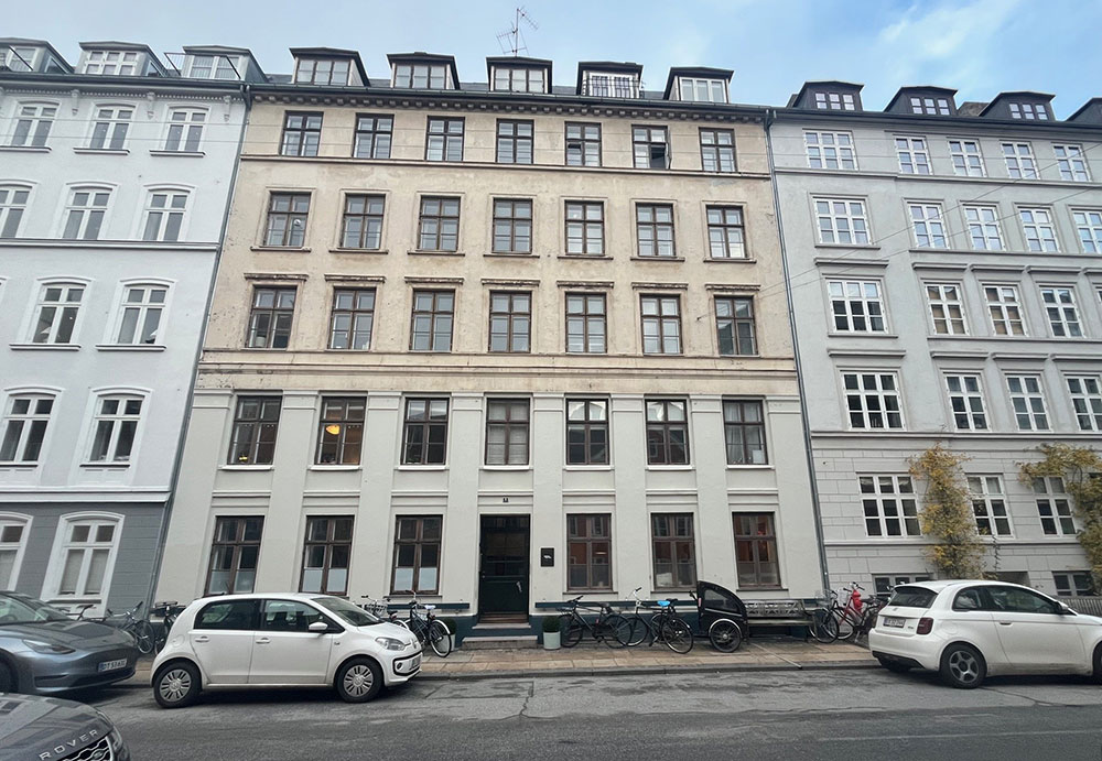 Lejlighedsfacade set fra gadeplan. Foran lejlighed bygningen ses biler og parkerede cykler.