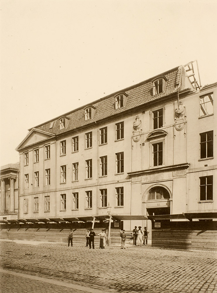 Sort/hvid billede af en bygningsfacade i nyklassicistisk stil. Foran bygningen ses et træhegn og en gruppe mænd med arbejdsredskaber.