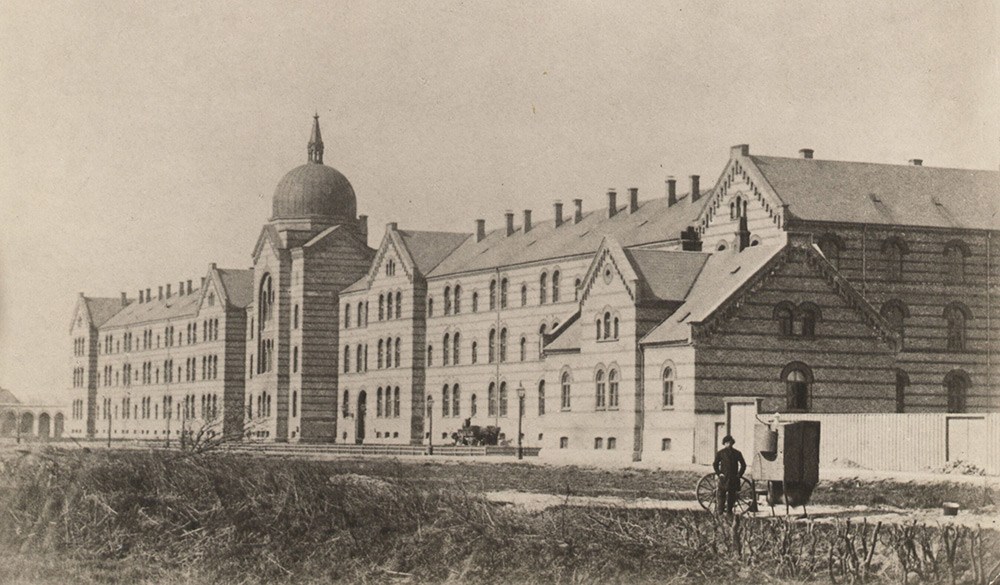 Sort/hvid billede af en stor treetagers bygning med kuppel i midten. Bygningens facade ses skråt fra højre mod venstre. I forgrunden ses en mand med en vogn.