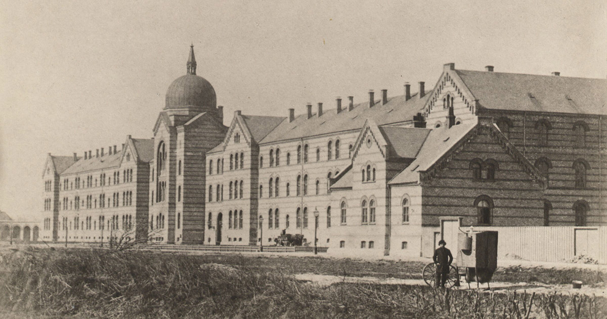 Sort/hvid billede af en stor treetagers bygning med kuppel i midten. Bygningens facade ses skråt fra højre mod venstre. I forgrunden ses en mand med en vogn.
