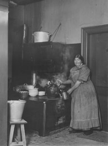 En kvinde med forklæde står i et køkken. Kvinden hælder dampende vand fra en kedel på komfuret ned i en kande. I køkkenet ses også baljer og bunker af service.